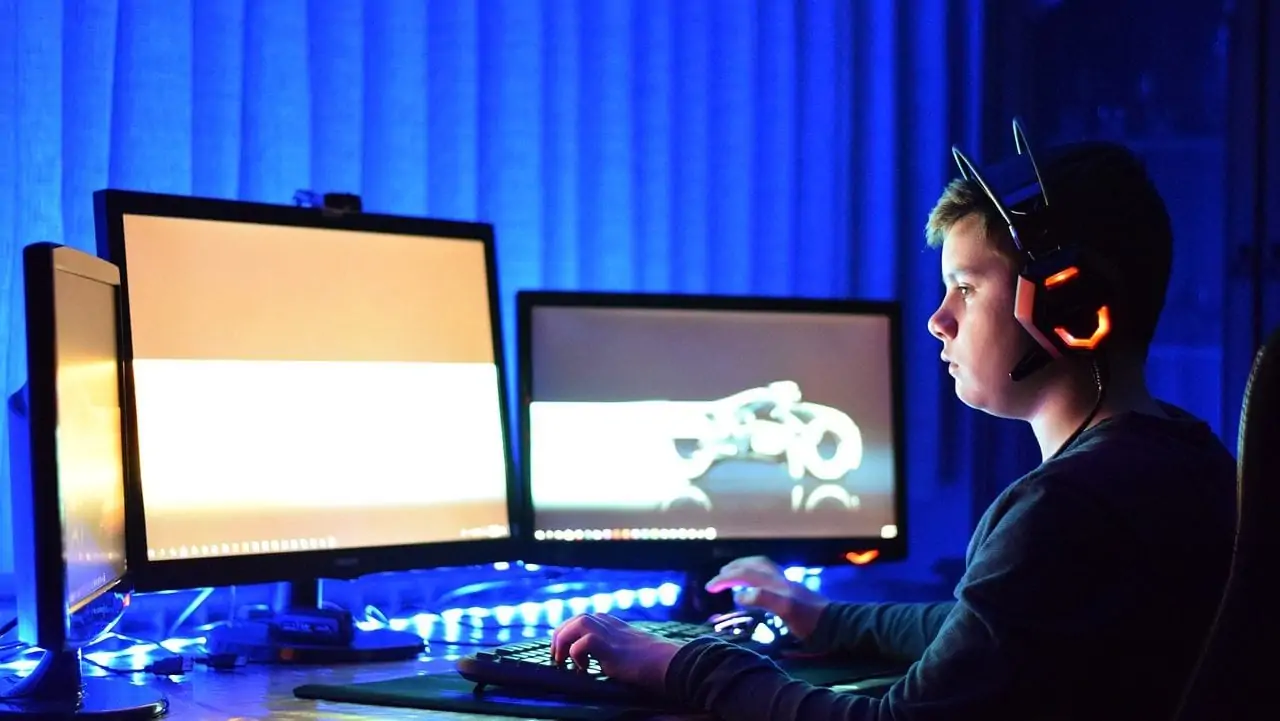 Jongen die aan het gamen is op drie beeldschermen, met headset op. Zij-aanzicht