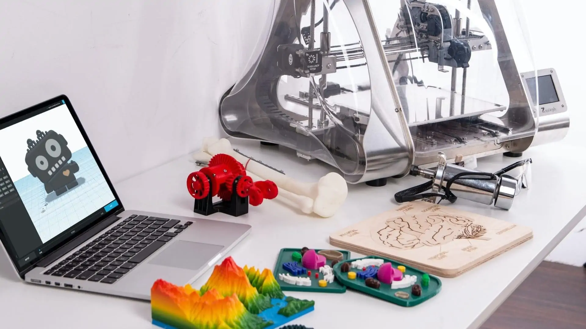 3D-printer naast laptop met allerlei prints, vooraanzicht
