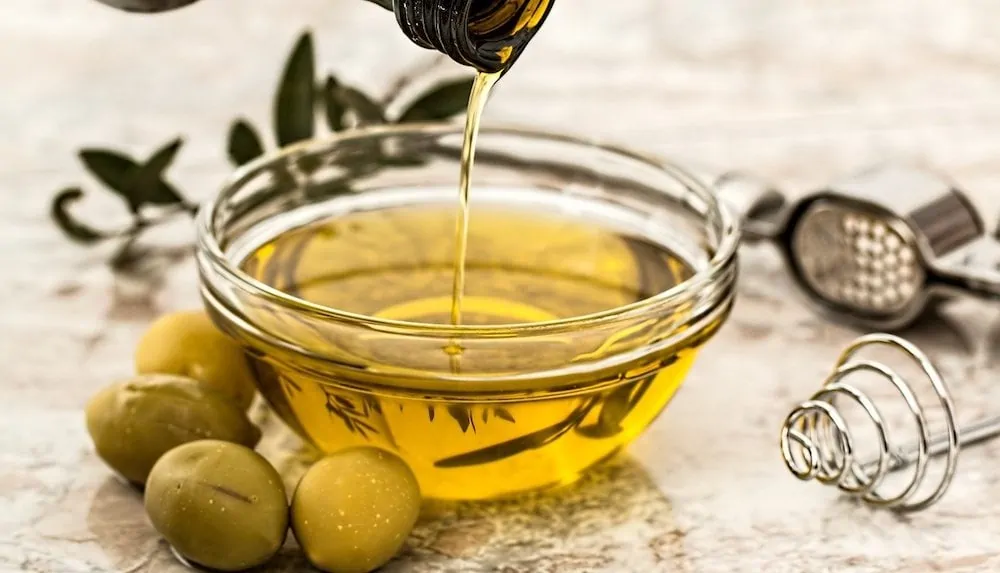 Glazen kommetje waar olijfolie in wordt gegoten met olijven eromheen.