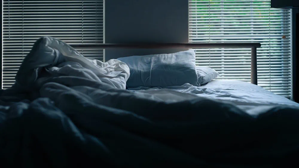 Afbeelding van een onopgemaakt bed voor twee ramen met jaloezieën. 