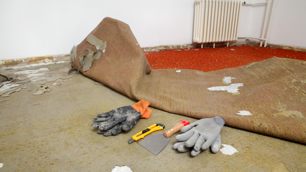 Twee paar werkhandschoenen, een verfkrabber en een stanleymesje liggen op de grond voor verwijderd tapijt