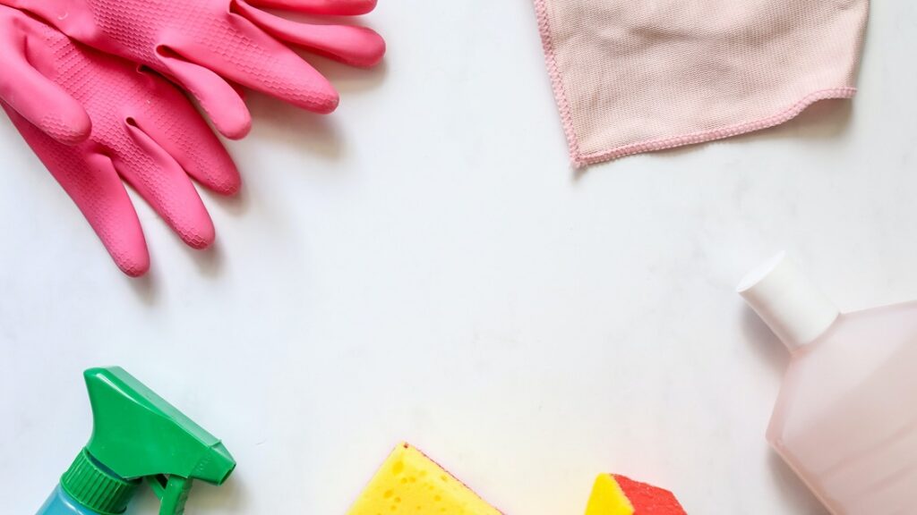 Knalroze plastic handschoenen, licht roze microvezeldoekje, groen sprayflesje, geel sponsjes en een fles.