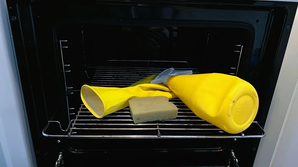 Een schoonmaakhandschoen, spons en spuitfles op het rooster van een geopende oven.